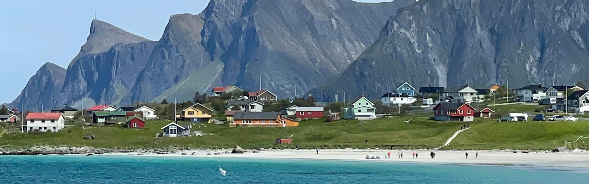 En strand med hav,  fjell og bebyggelse i ulike farger, i bakgrunnen. Foto