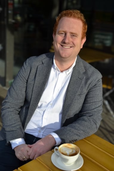 En person med rødt hår og grå blazer sitter på en uteservering med en kaffe foran seg og smiler til kamera.
