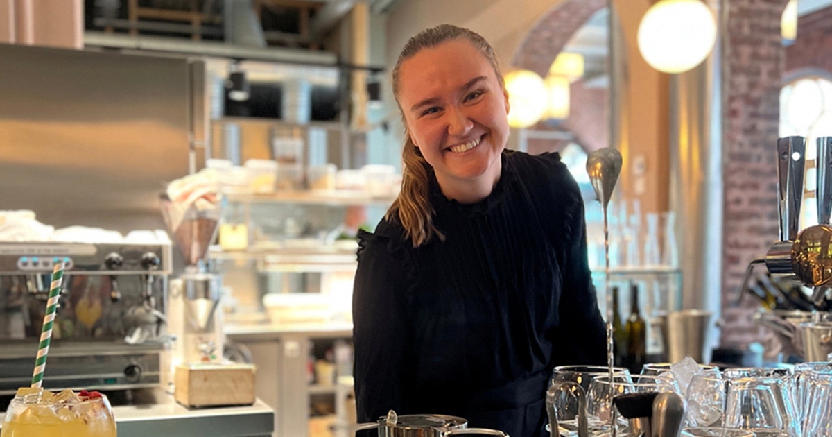 en ung, smilende kvinne som står bak en bar. bak henne sees en kaffemaskin i stål. 