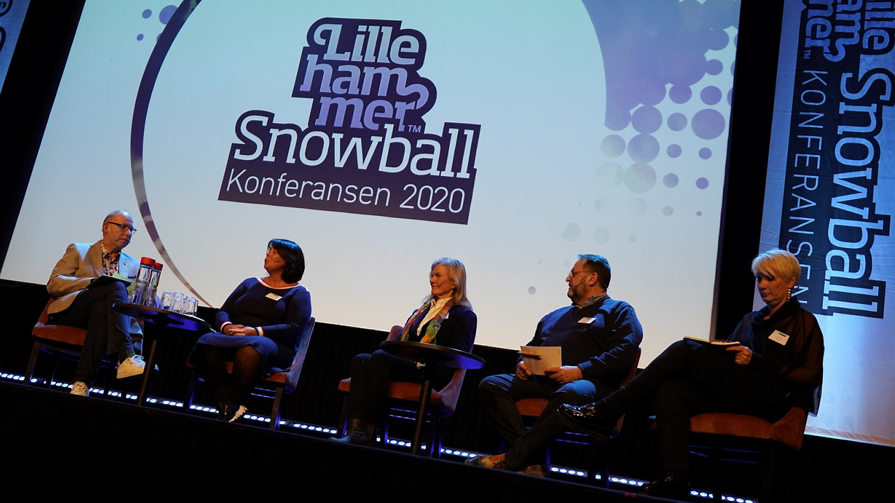 Bilde tatt nedenfra opp mot en scene der fire paneldeltagere sitter og ser mot moderator. I bakgrunnen er en hvit skjerm med teksten Snowball-konferansen.