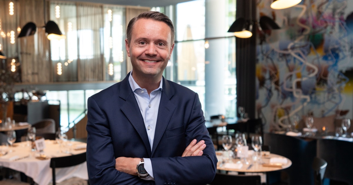 En smilende mann i dress som står i et restaurantlokale. Foto.