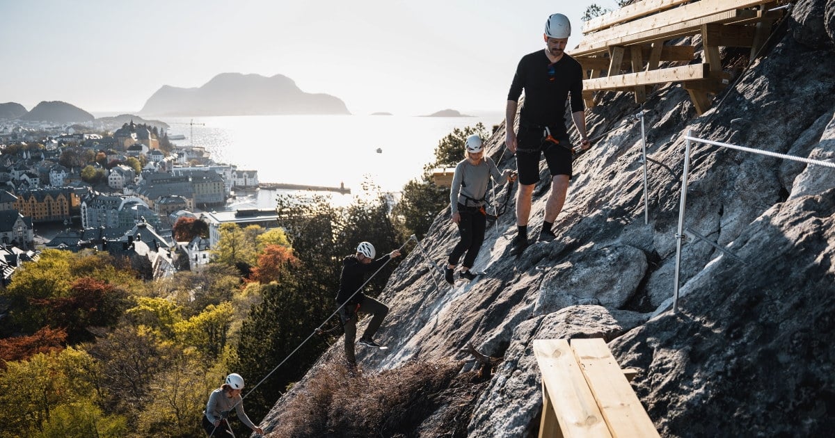 En gruppe personer med hjelm og klatreutstyr beveger seg langs en via ferrata-rute i fjellsiden over Ålesund, med byen, havet og øyene i bakgrunnen.