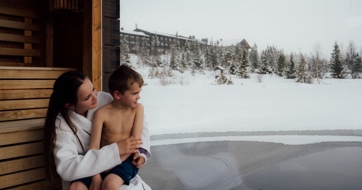 En kvinne og et barn sitter i en badstue og ser ut på et vinterlandskap med snø, trær og et hotell i bakgrunnen.