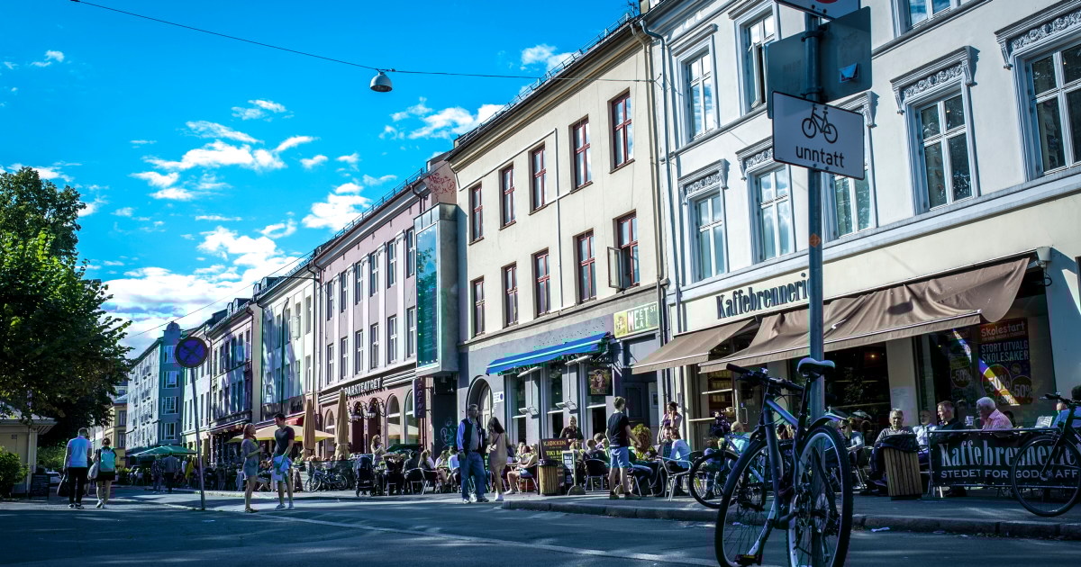 Gatebilde fra Grünerløkka i Oslo med uteservering og mennesker som nyter mat og drikke i sola. Fortauskafeer og små butikker ligger tett i tett langs bygårdene, og sykler er parkert langs fortauskanten. En blå himmel med noen skyer i bakgrunnen.