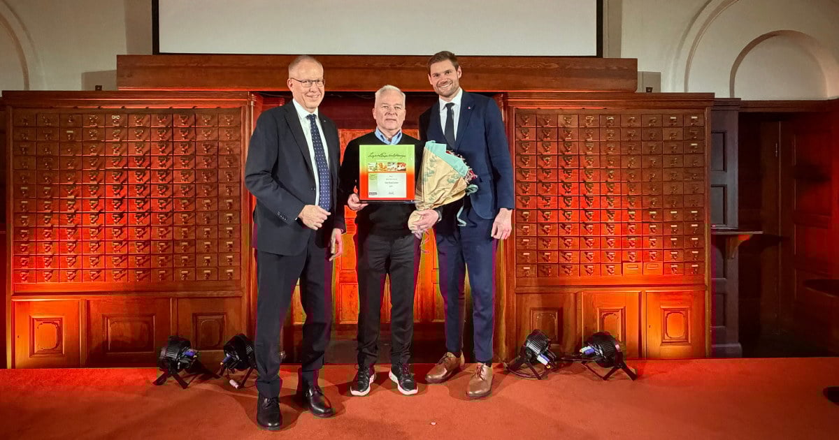 tre menn som står på en scene. Den ene holder en plakett og en blomsterbukett. Foto