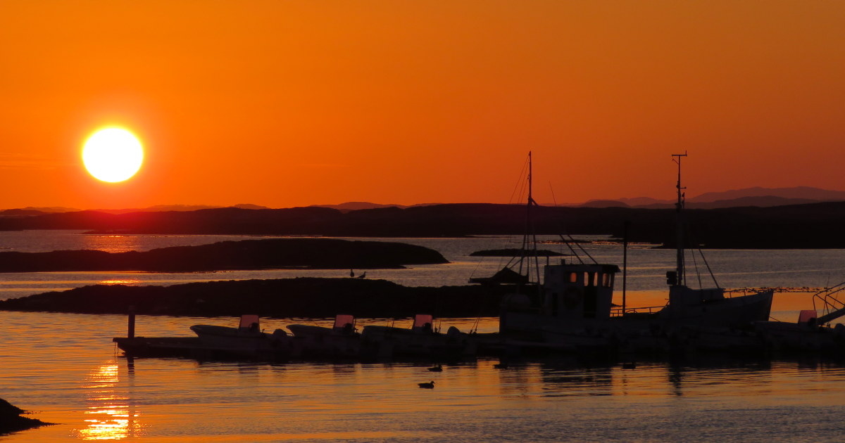 Silhuetter av båter og øyer mot en oransje himmel ved solnedgang. Foto.