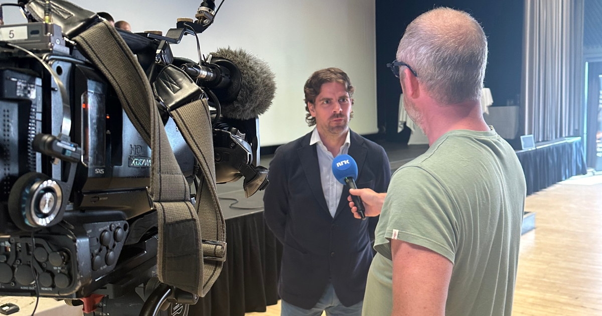 Mann kledd i dresjakke som intervjues av en mann i grønn t-skjorte med et TV-kamera i forgrunnen.