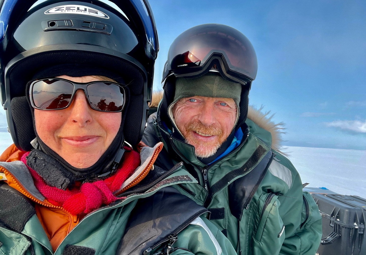 To personer i varme vinterklær og hjelmer smiler mot kamera ute på snødekt landskap. Den ene har solbriller og rosa skjerf, den andre har hjelm med solbeskytter dradd opp over pannen. Klar blå himmel i bakgrunnen.