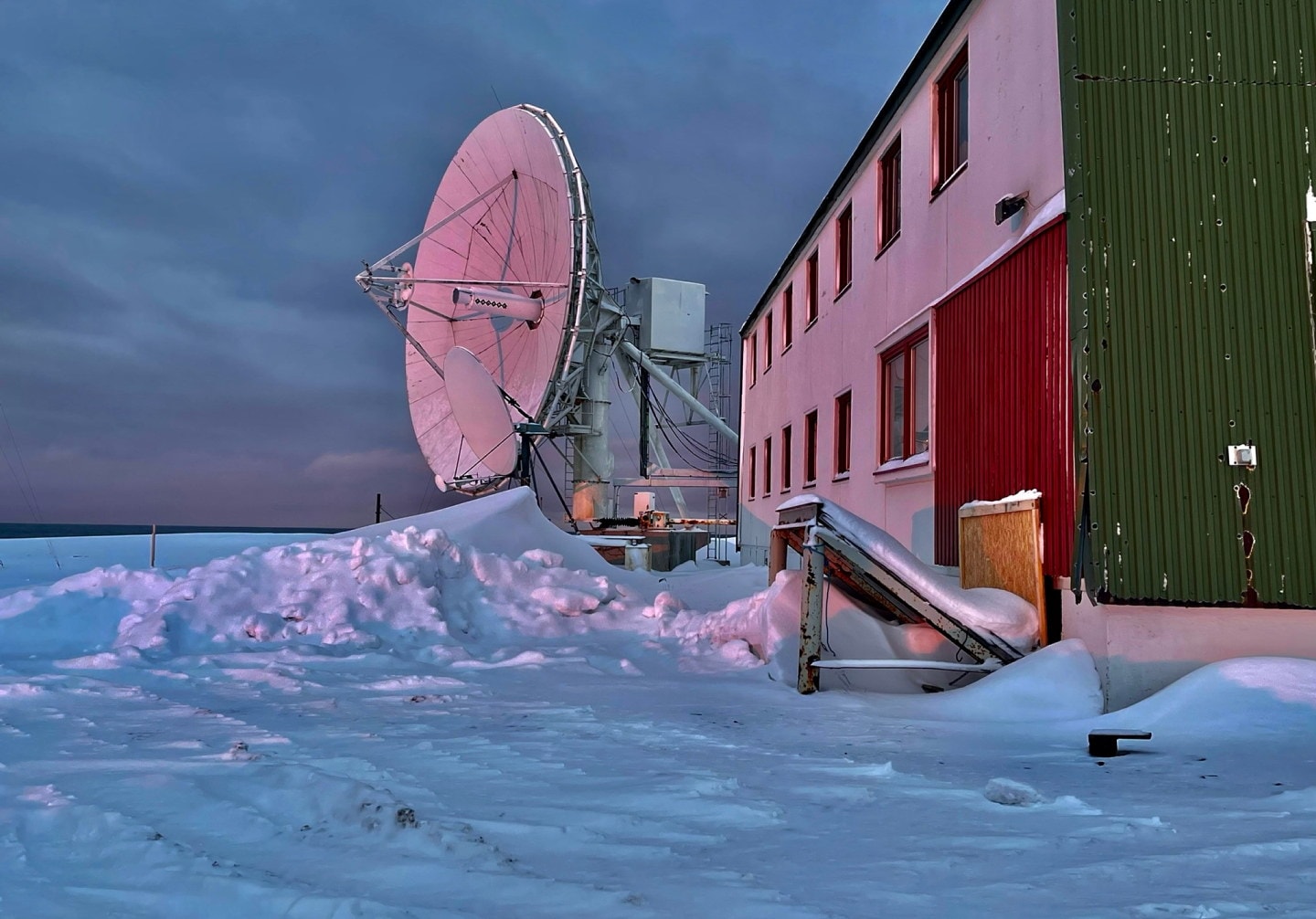 Stor satellittantenne ved siden av en bygning, omgitt av snø, med vakker himmel i bakgrunnen.