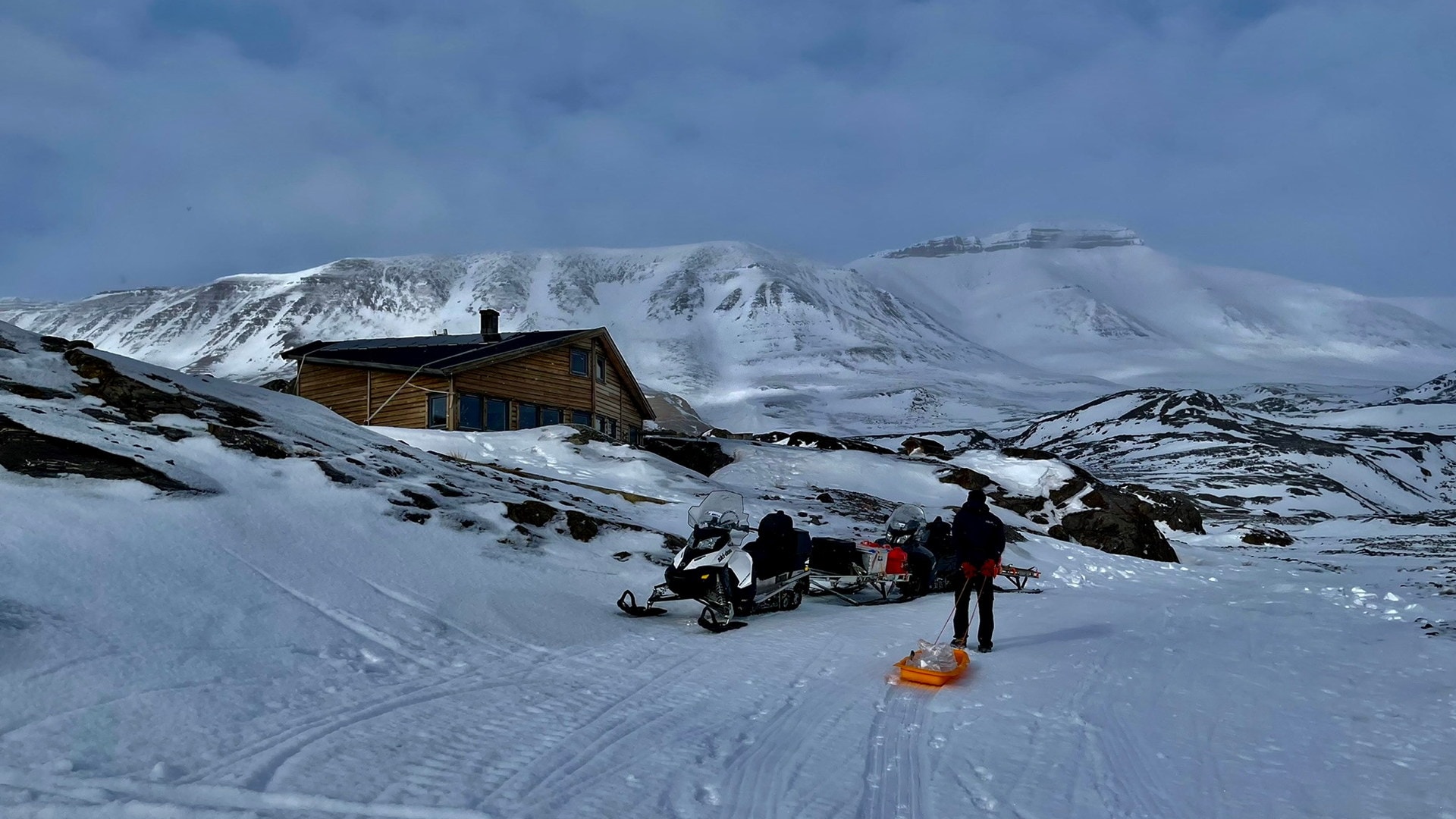 Person trekker en pulk i snødekt landskap mot en hytte omgitt av fjell. En snøscooter med utstyr står parkert i snøen.
