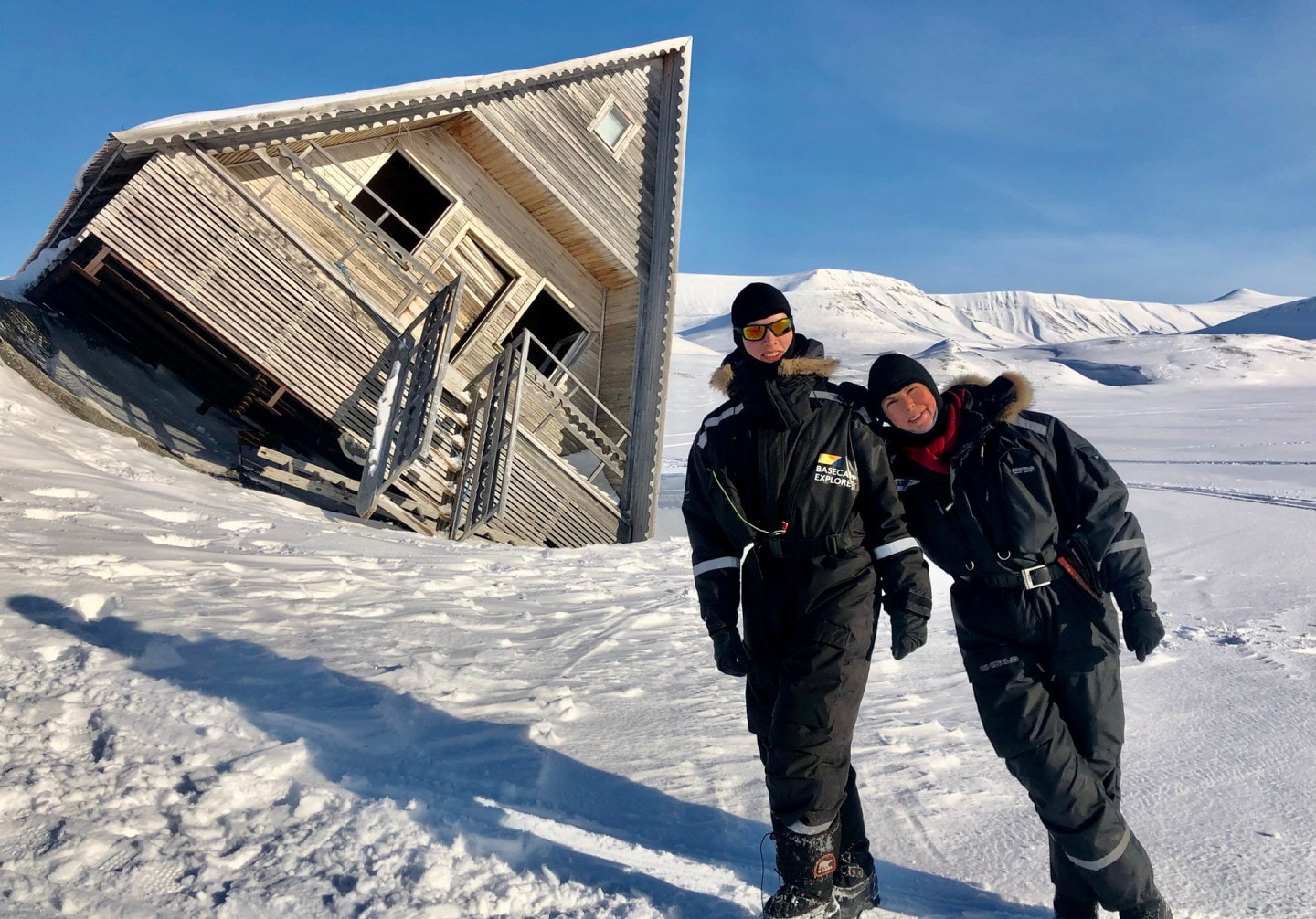 To personer i varme vinterdresser står i snøen foran et trehus som har sunket ned i snøen på den ene siden, og nå ser helt skjevt ut. Snødekte fjell og blå himmel i bakgrunnen.