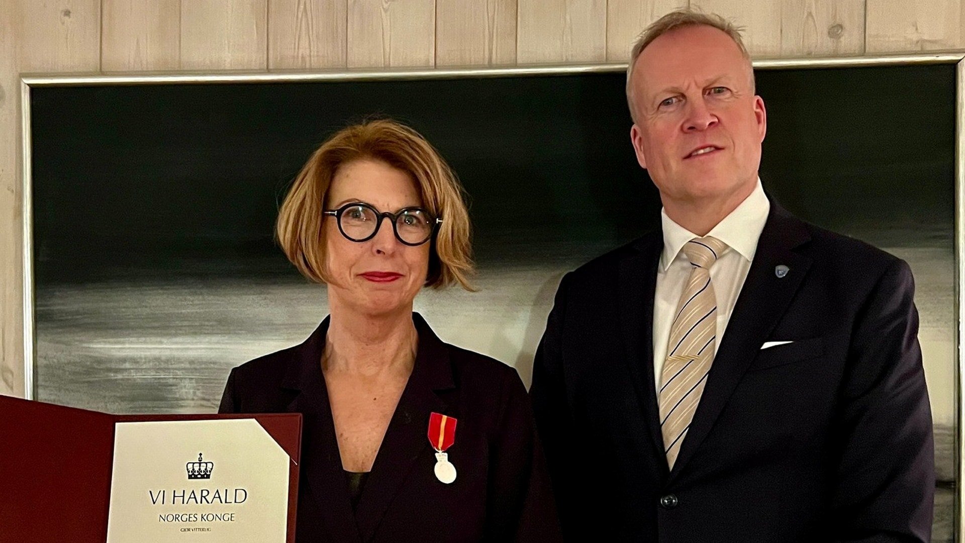Bilde av en kvinne og en mann i formelle klær. Kvinnen holder et diplom med kongens navn og bærer Kongens fortjenstmedalje på jakken. Mannen står ved siden av henne med hendene samlet foran seg.