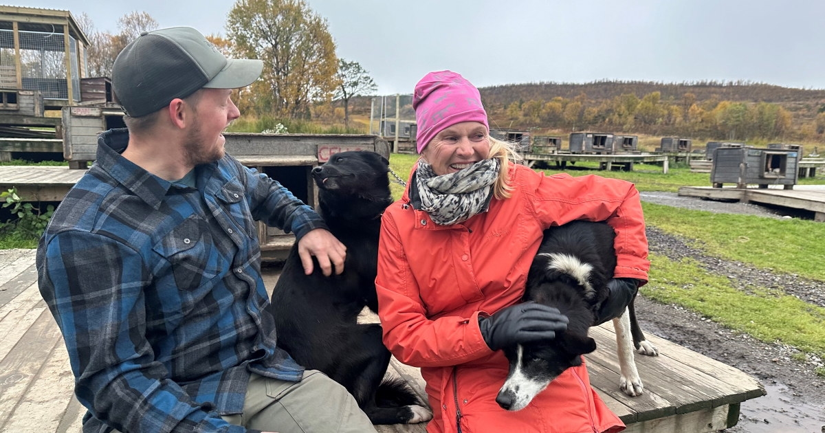 Kvinne og mann koser med to hunder foran en hundegård.