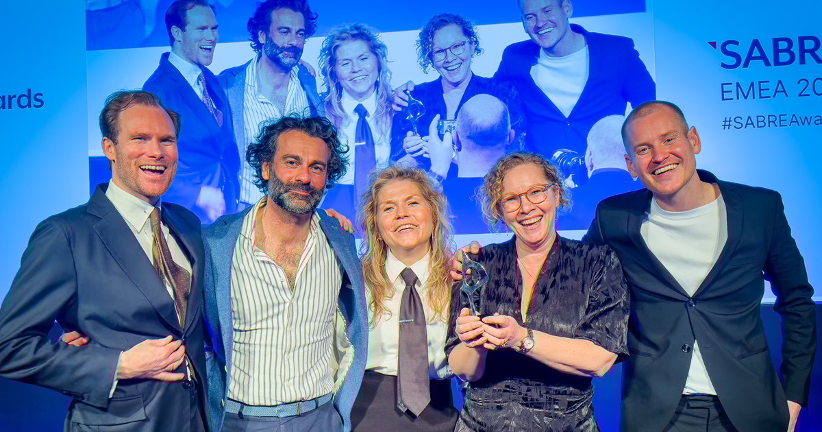 Fem glade personer står samlet på en scene med en pris i hånden, foran en skjerm som viser et bilde av dem i bakgrunnen. De smiler og feirer seieren under SABRE Awards EMEA.