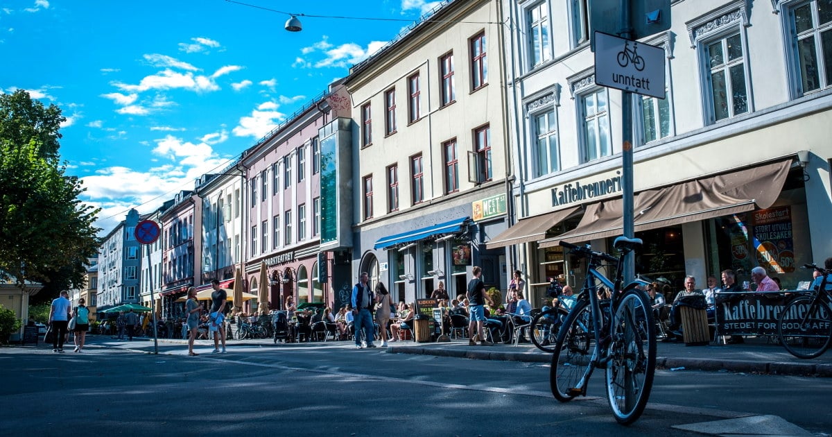 Gatebilde fra Grünerløkka i Oslo med uteservering og mennesker som nyter mat og drikke i sola. Fortauskafeer og små butikker ligger tett i tett langs bygårdene, og sykler er parkert langs fortauskanten. En blå himmel med noen skyer i bakgrunnen.