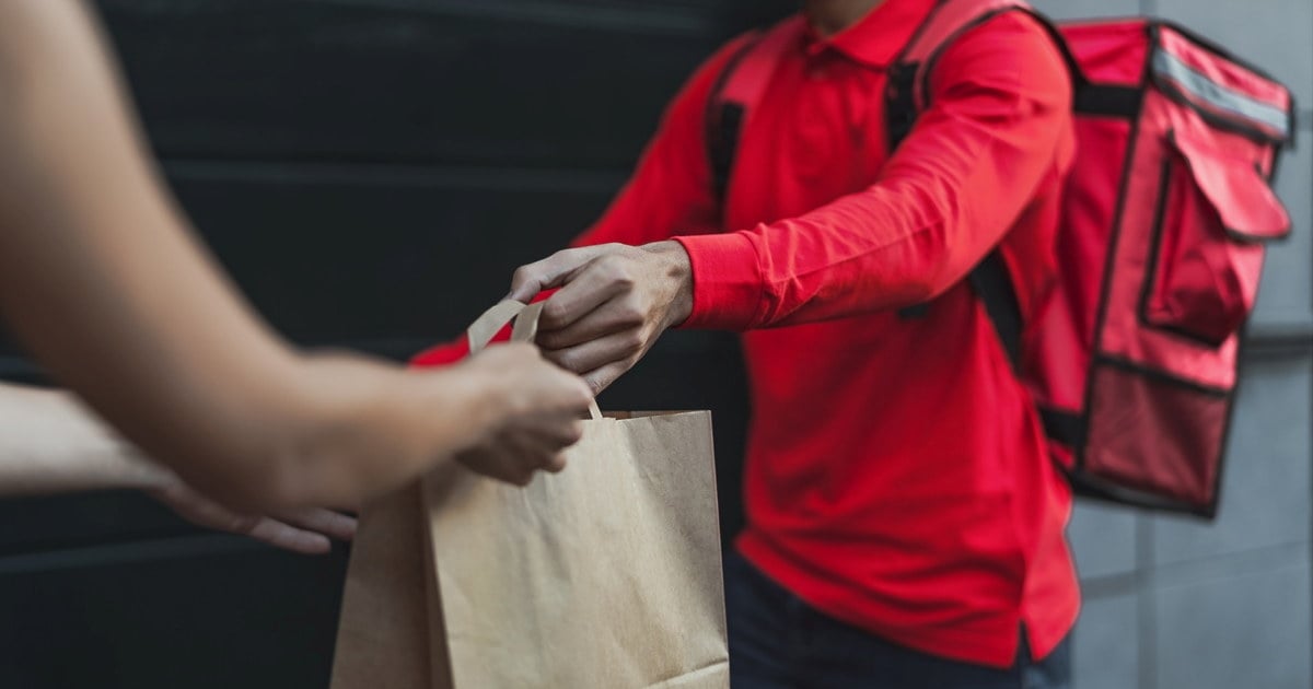 En budleverandør i rød uniform og med rød leveringsekk over skulderen overrekker en papirpose med mat til en kunde. Bildet viser kun hendene og overkroppene til personene. Bakgrunnen er en mørk vegg.