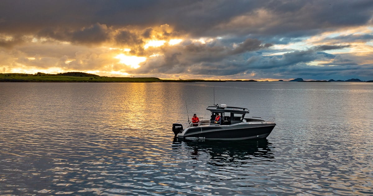 En småbåt med to personer i redningsvester ligger stille på sjøen i solnedgang. Det er lavtliggende land i bakgrunnen og dramatisk skydekke. Foto