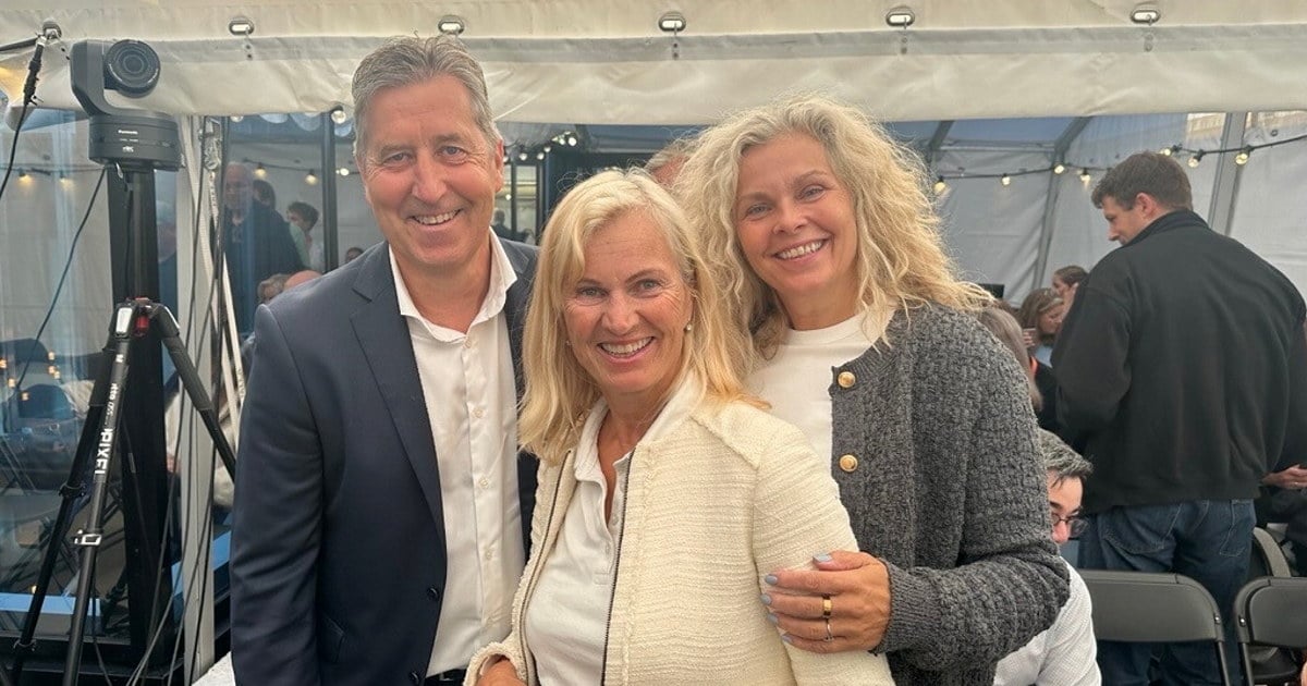 Tre personer står tett sammen og smiler til kamera i et lyst telt med mange mennesker i bakgrunnen. Det er mikrofon- og kamerautstyr synlig til venstre i bildet. Foto.