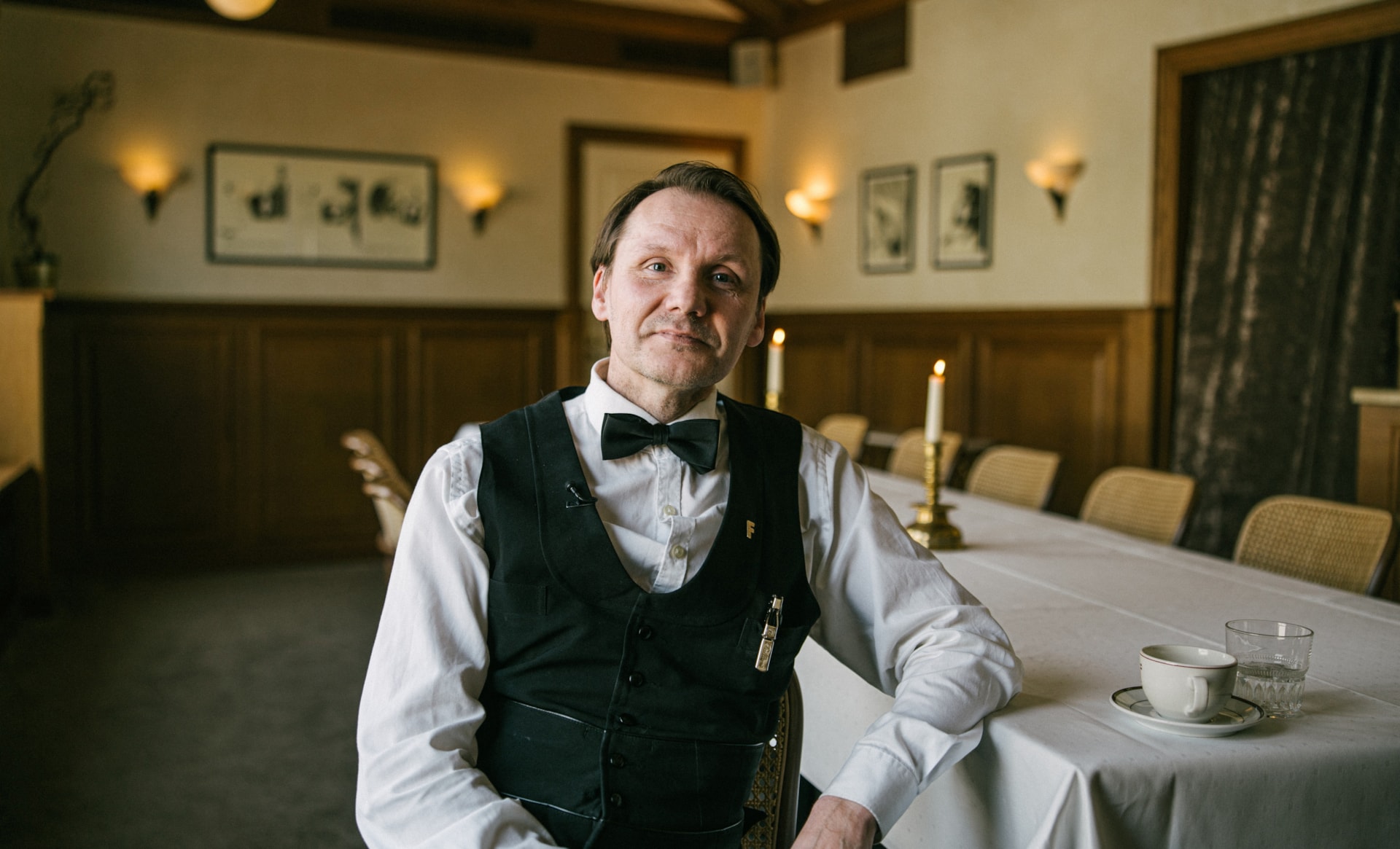 Servitør i hvit skjorte, sort vest og sløyfe sitter ved et dekket langbord i et klassisk innredet rom med tente lys.