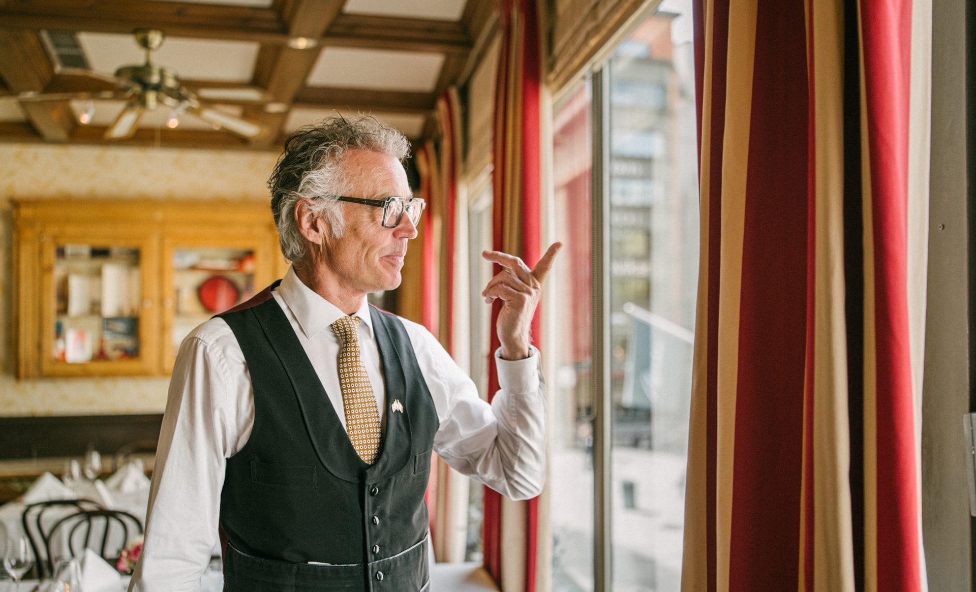 Mann i hvit skjorte, slips og svart vest står inne i en restaurant med røde og gule gardiner og klassisk innredning. mannen ser ut av vinduet og peker ut. Foto