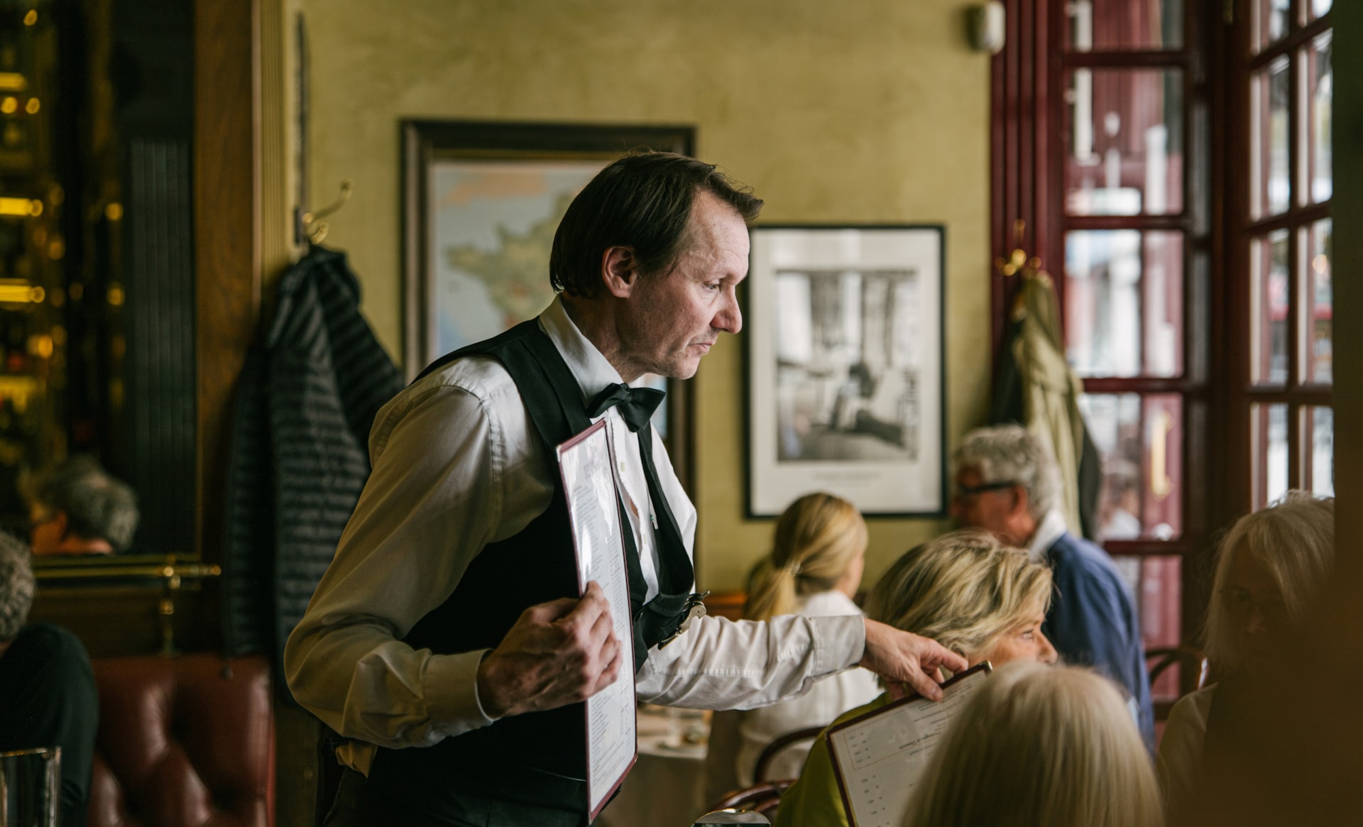 Servitør i hvit skjorte, sort vest og sløyfe holder menyer og snakker med gjester ved et bord i en travel restaurant. Veggen bak har innrammede bilder og vinduer med røde sprosser.