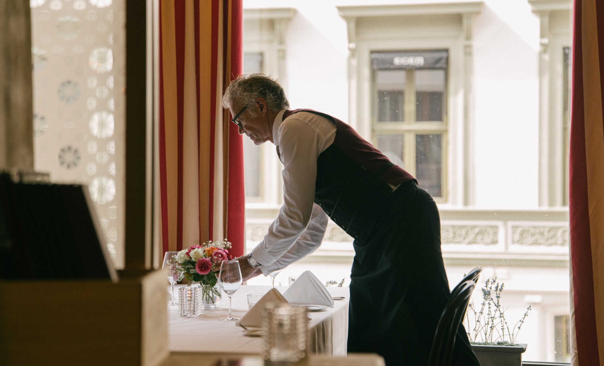 En servitør dekker et restaurantbord med hvit duk, servietter og glass, mens han bøyer seg over bordet i et lokale med store vinduer og røde gardiner.