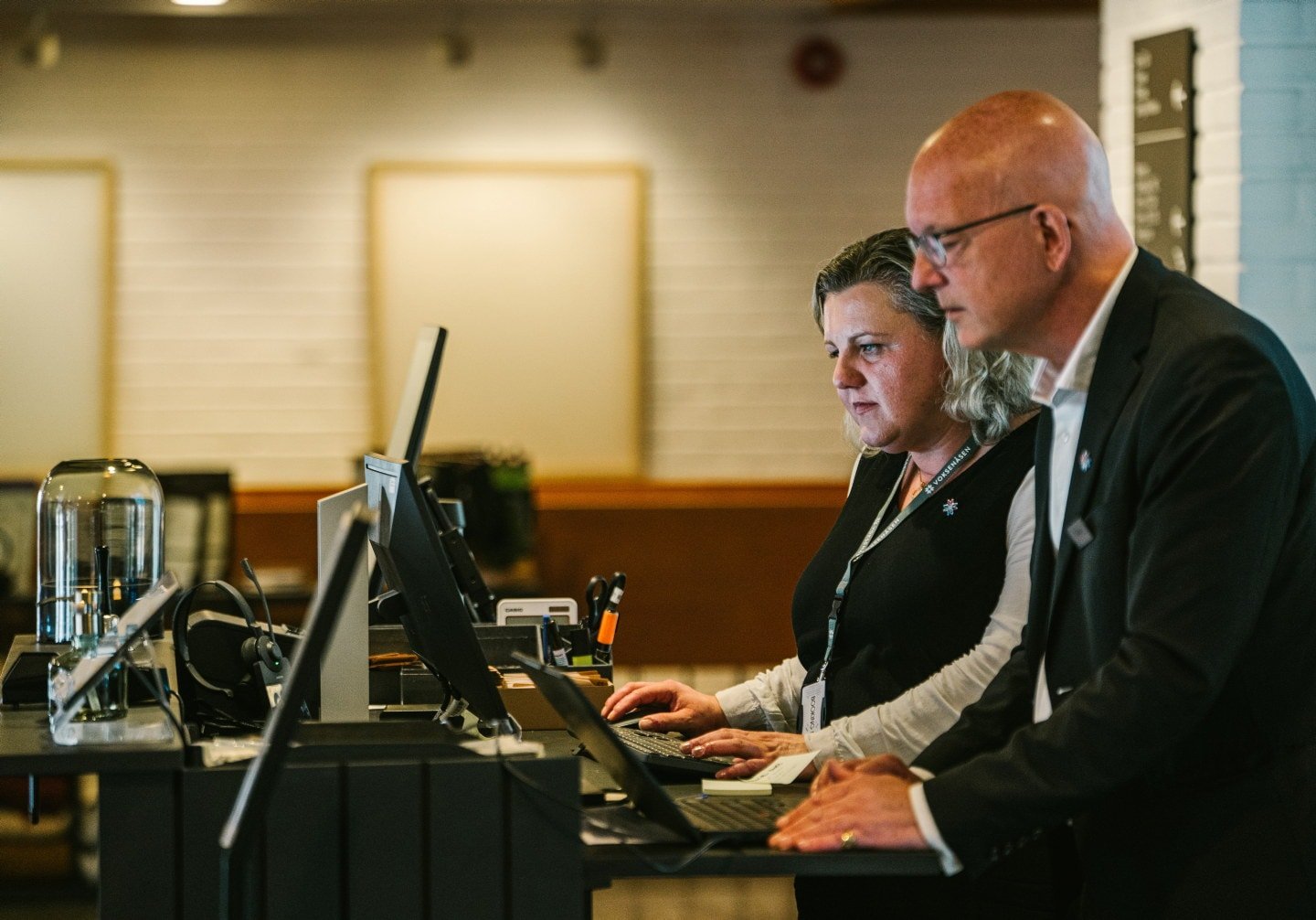 To ansatte bak resepsjonen på et hotell. Kvinnen har et nøkkelkort i snor rundt halsen og står ved en datamaskin, mens mannen ved siden av har et navneskilt festet på jakkeslaget.