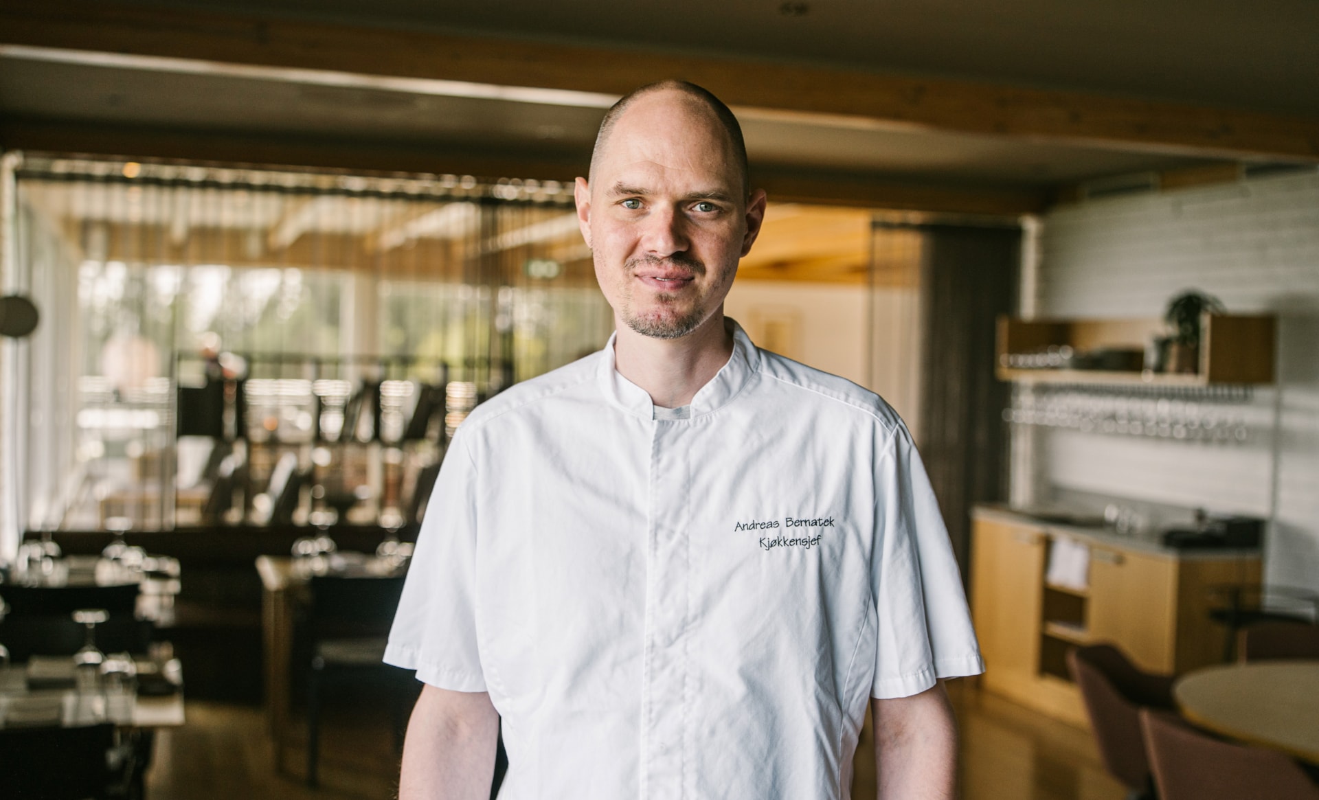 Kjøkkensjef i kokkeklær står midt i en moderne restaurant og smiler mot kameraet. Navnet og tittelen "Kjøkkensjef" er brodert på kokkejakken.