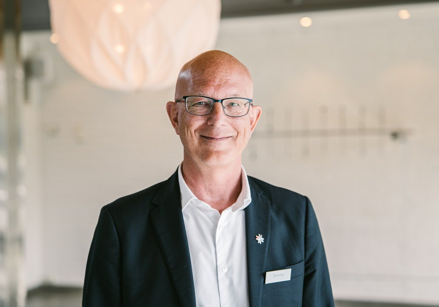 Mann med navneskilt og dressjakke smiler til kamera i et lyst og moderne rom med hvit vegg og hengende lampe i bakgrunnen.