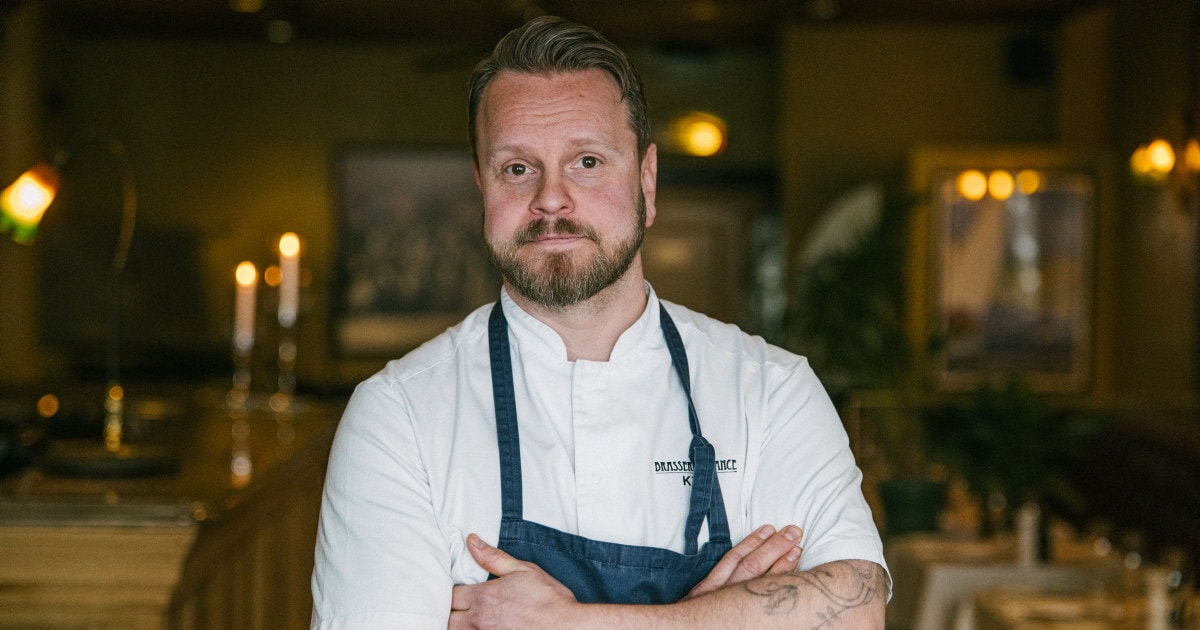 En mann i kokkeklær og mørkeblått forkle står med armene i kors og et alvorlig uttrykk i en stemningsfull restaurant.