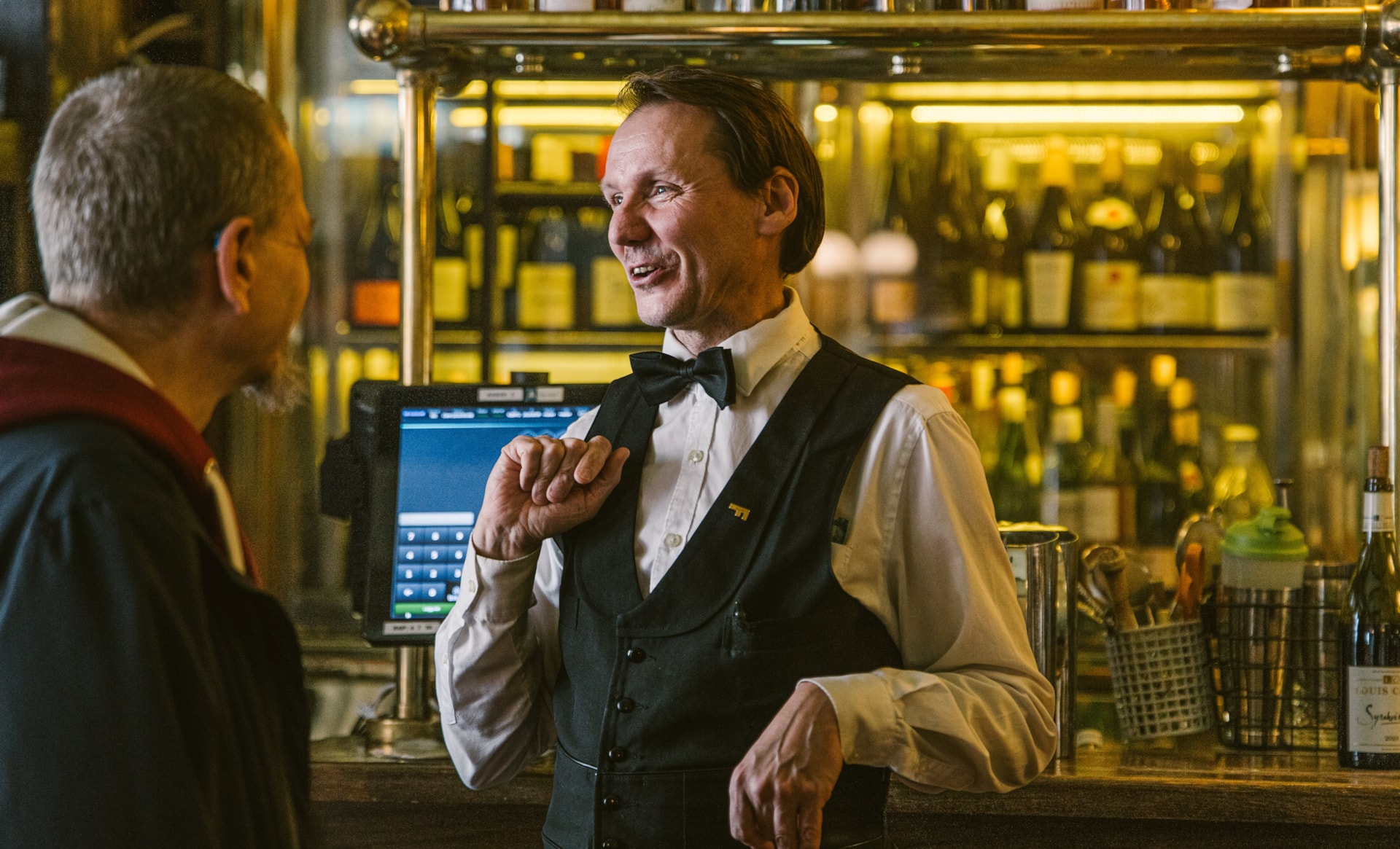 ervitør i uniform står bak baren og prater med en gjest, med vinflasker og glass i bakgrunnen.