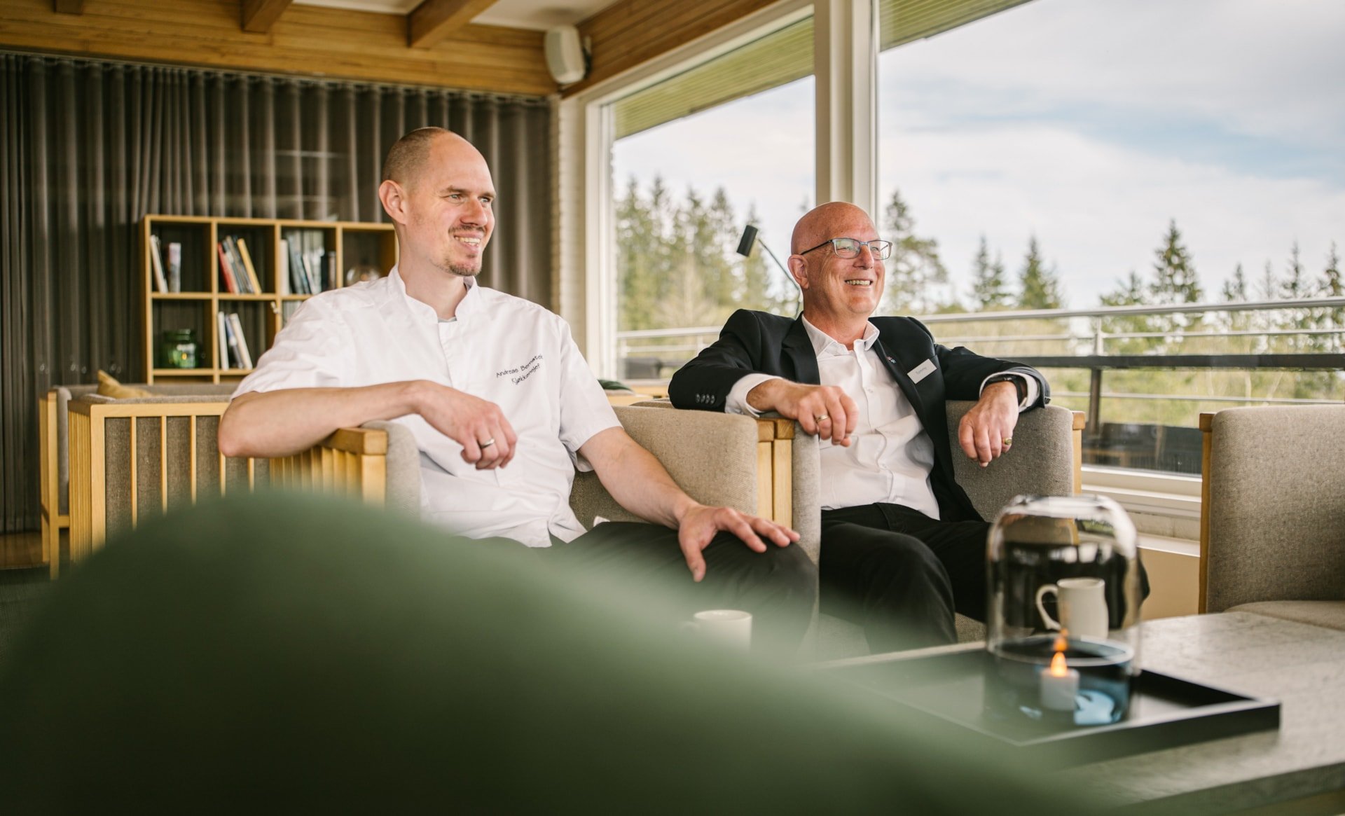 To menn sitter i en lounge med store vinduer og utsikt mot skog. Den ene er iført kokkeklær, den andre har på seg dressjakke med navneskilt. På bordet foran dem står en kanne med kaffe og en kopp.