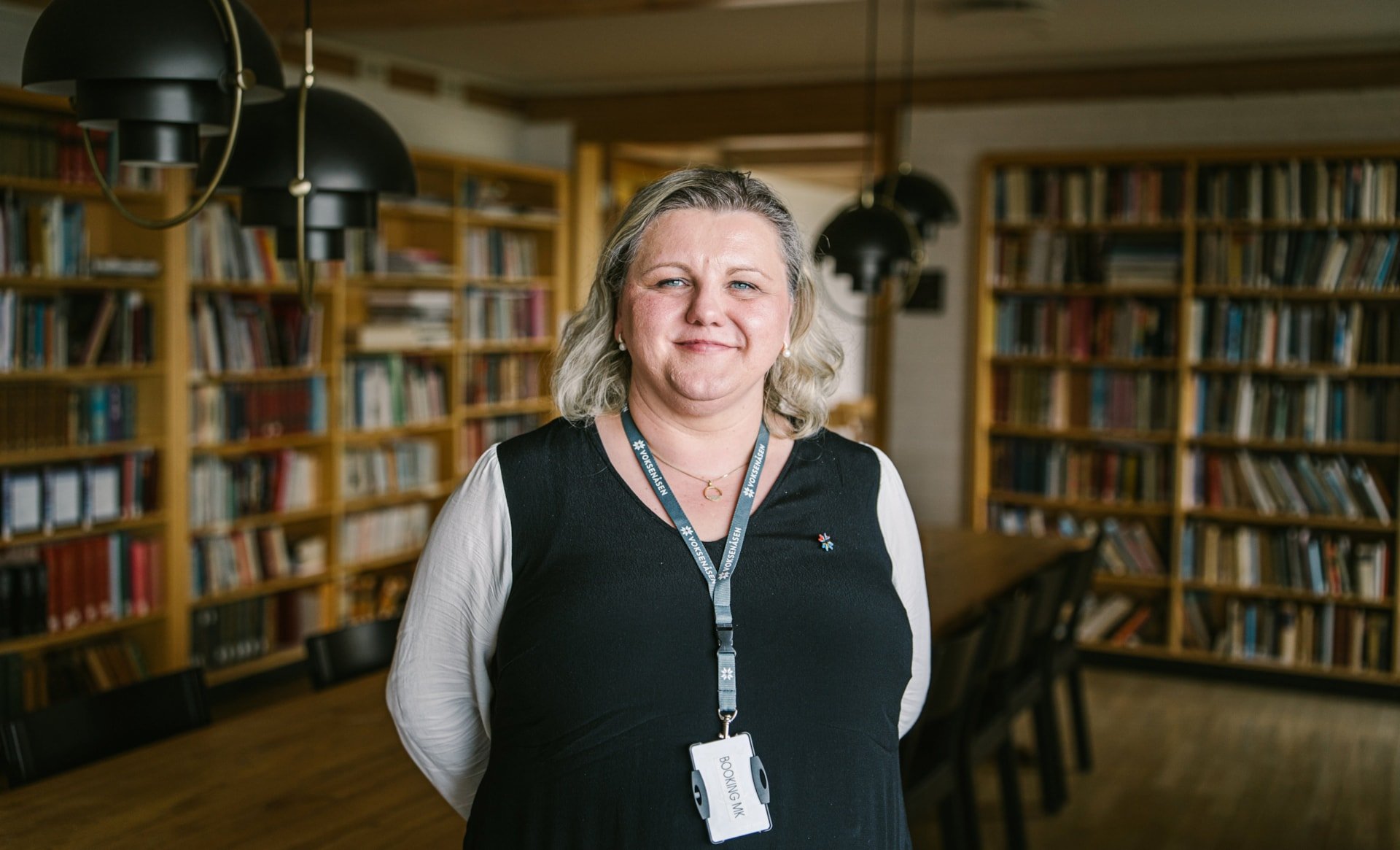 En kvinne står i et bibliotek med bokhyller langs veggene. Hun har på seg et mørkt antrekk med lys langermet skjorte under, og et nøkkelkort i snor rundt halsen merket «Booking MK». Hun smiler og ser rett mot kamera.