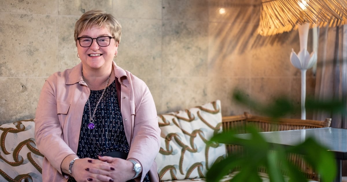 Portrettfoto av en smilende kvinne som sitter med hendene foldet, i en mønstret sofa. En plante sees i forgrunnen. Foto.