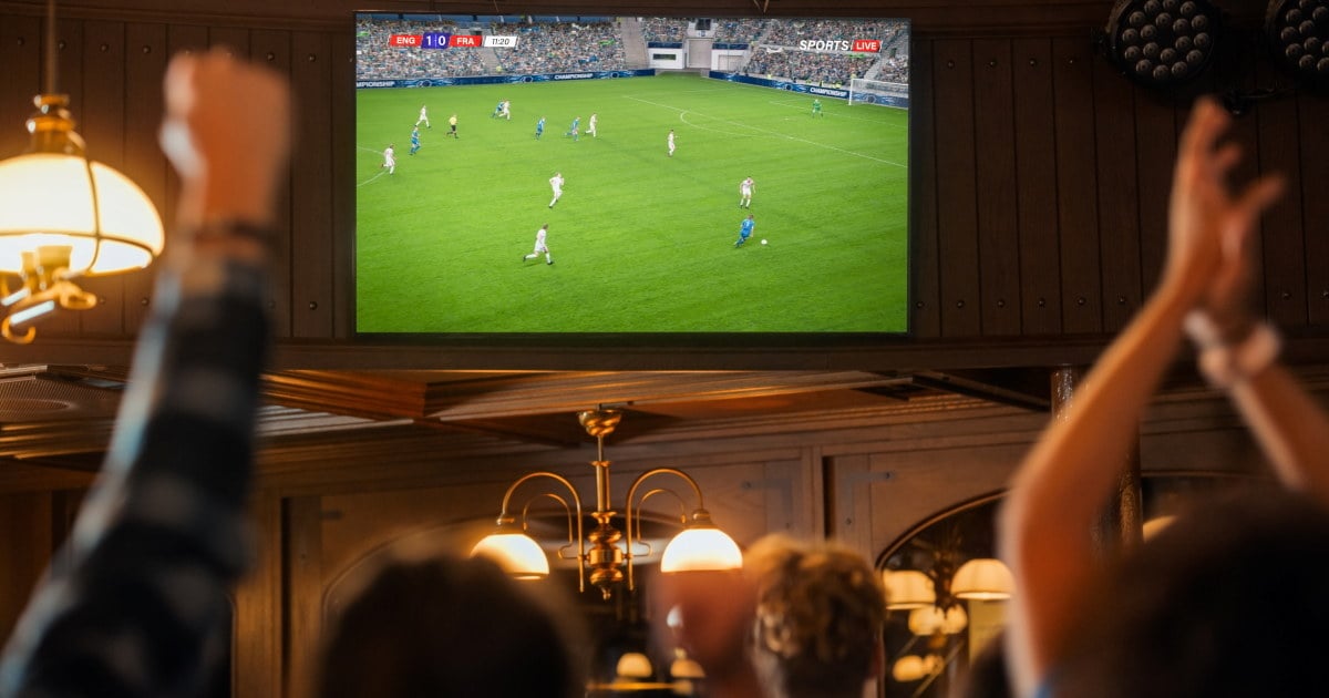 Storskjerm med direktesendt fotballkamp i fokus i en pub, mens mennesker i forgrunnen jubler med armene i været.