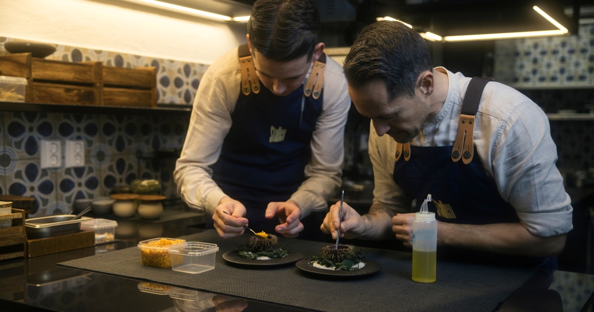 To kokker i forklær arbeider konsentrert side om side ved kjøkkenbenken og finjusterer hver sin anrettede tallerken.