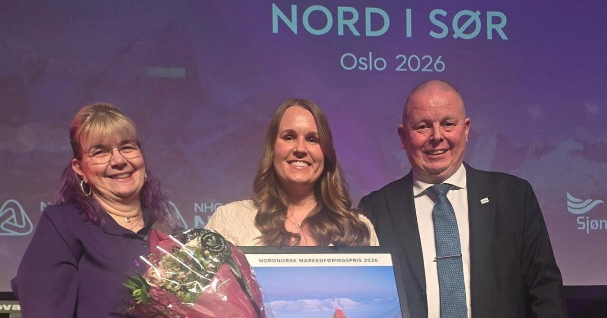 Tre smilende mennesker som står på en scene. Kvinnen i midten holder en blomsterbukett og en diplom.