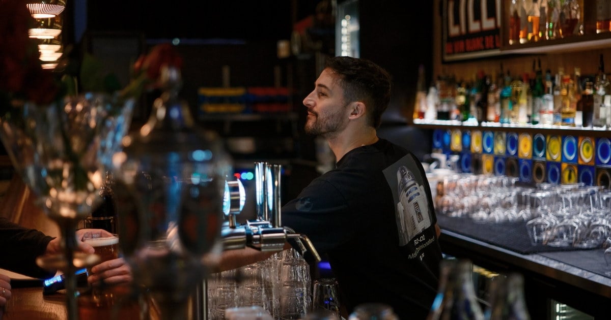 Person serverer drikke bak bardisk i restaurantlokale.