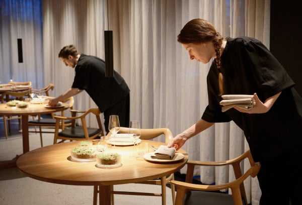 To ansatte klargjør bord med servietter, glass og dekketøy i et restaurantlokale før service.