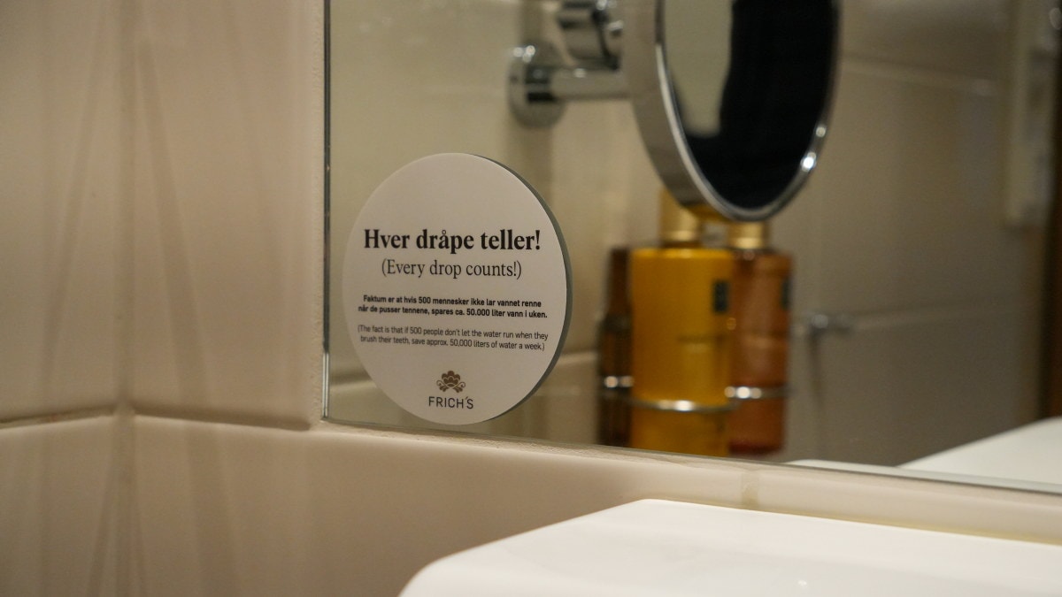 Skilt på et baderomsspeil med teksten «Hver dråpe teller!». En vask og et speil er synlige.