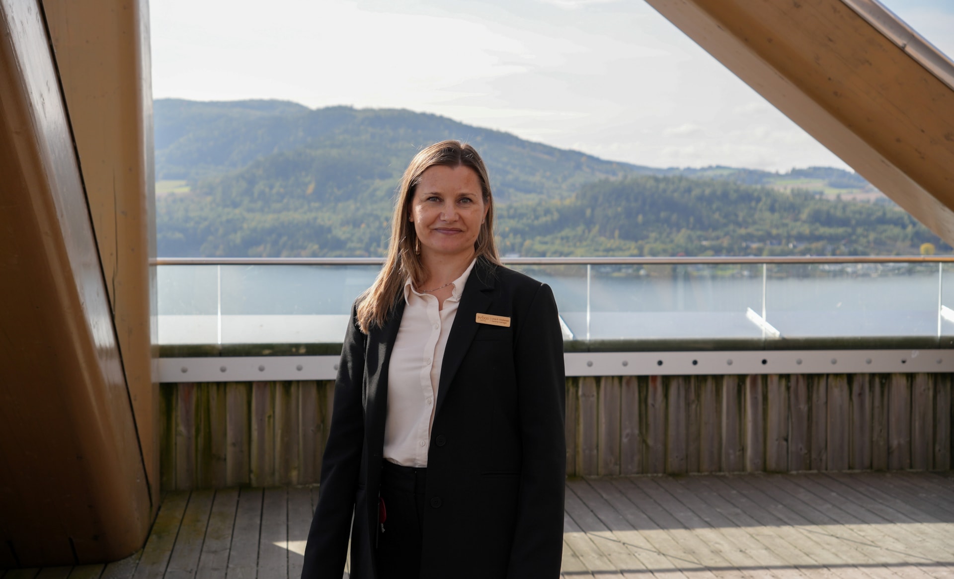 En person står på en terrasse foran glassrekkverk, vann og skogkledde åser.