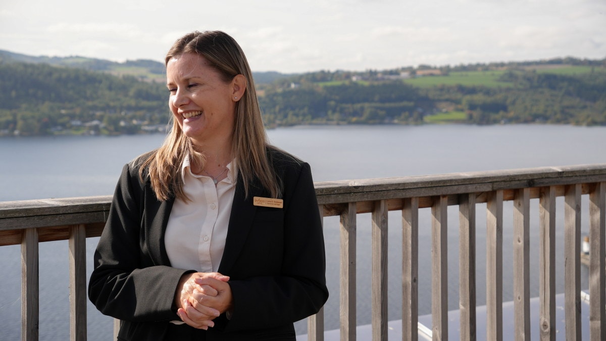 En person står ved et rekkverk og smiler. Vann og grønne åser er synlige i bakgrunnen.