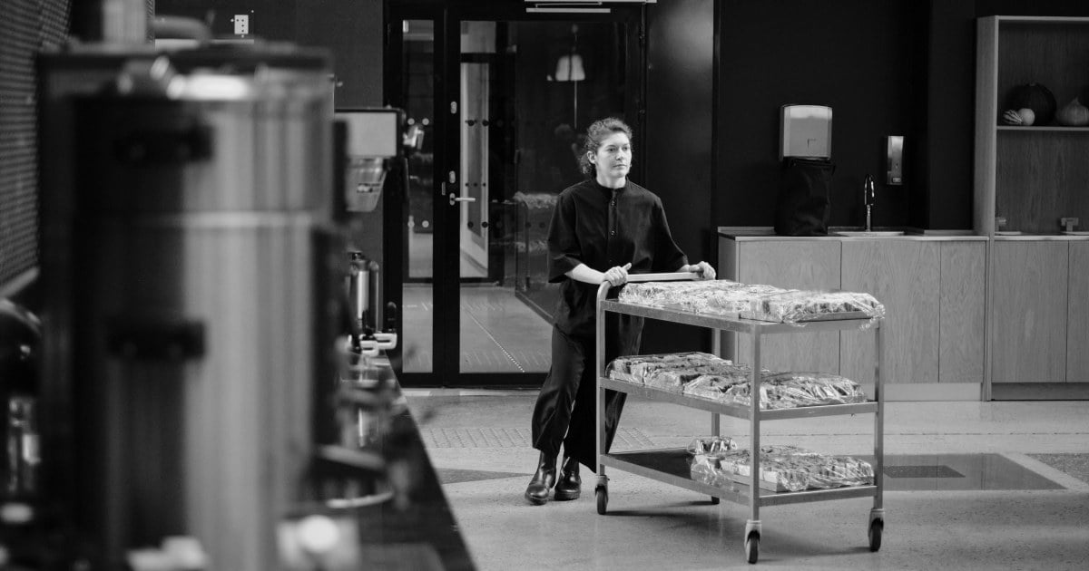 En ansatt skyver en tralle med ferdigpakket mat gjennom et profesjonelt kjøkkenområde med benker og utstyr i bakgrunnen.