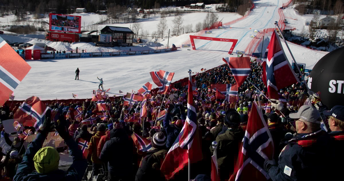 Publikum står tett i en tribune under et skirenn og vifter med norske flagg, med målområdet og løypa synlig i bakgrunnen.