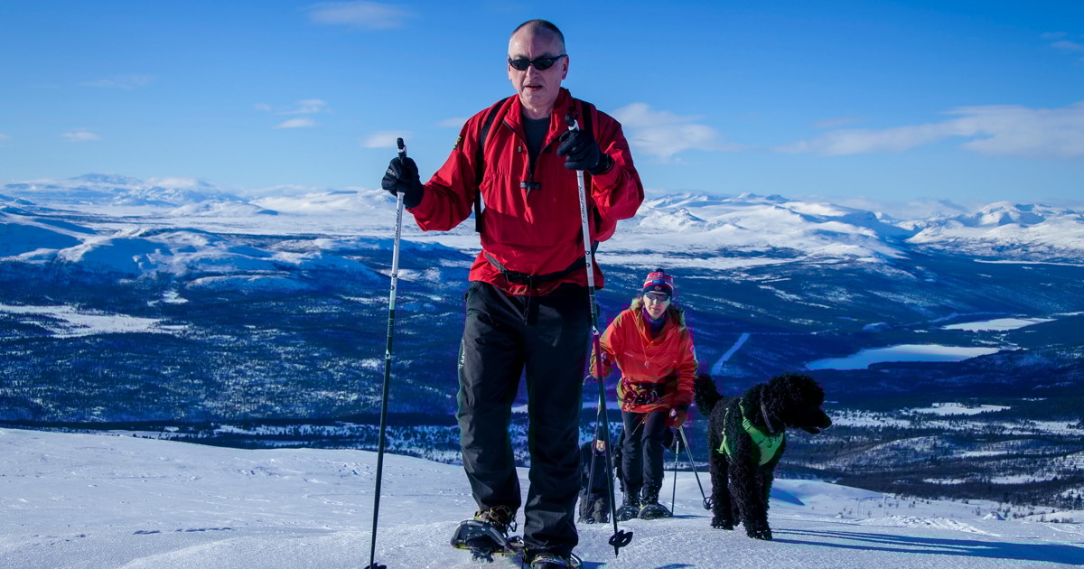 En mann, en kvinne og hund på trugetur opp et fjell med blå himmel i bakgrunn og masse snø rundt.