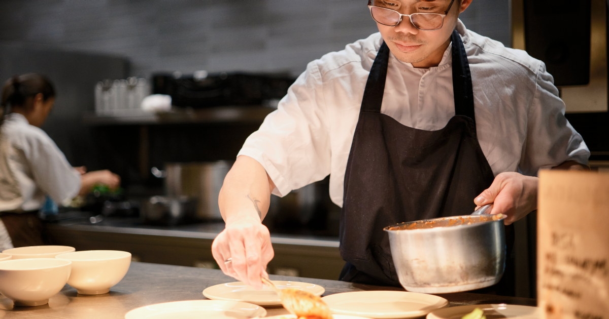 En kokk står ved kjøkkenbenken og anretter mat på tallerken med en kjele i hånden, mens et profesjonelt kjøkken er synlig i bakgrunnen.