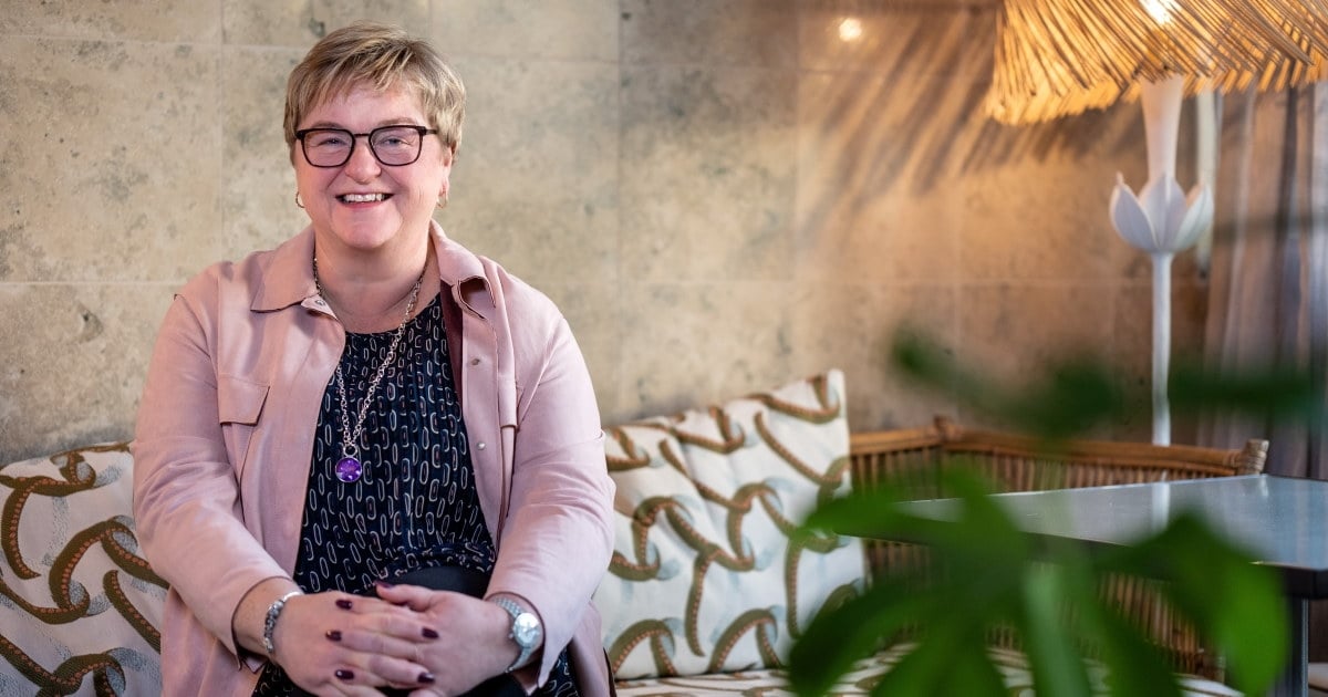 Portrettfoto av en smilende kvinne som sitter med hendene foldet, i en mønstret sofa. En plante sees i forgrunnen. Foto.