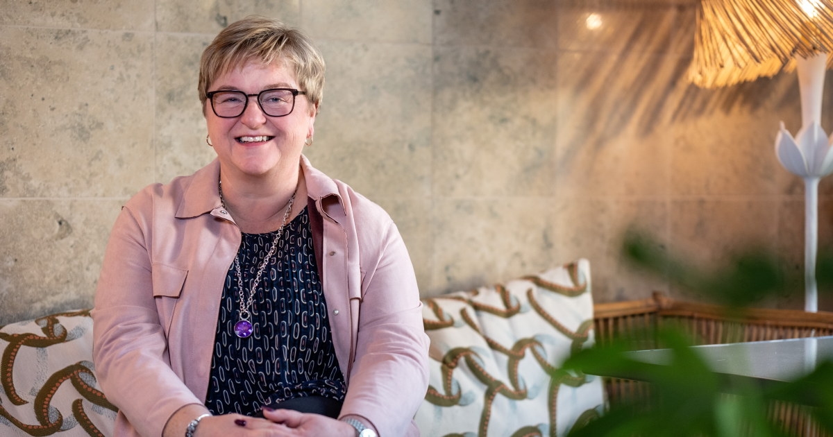 Portrettfoto av en smilende kvinne som sitter med hendene foldet, i en mønstret sofa. En plante sees i forgrunnen. Foto.