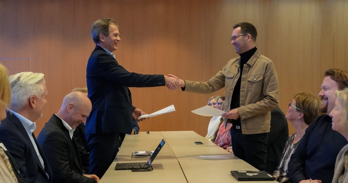 To personer håndhilser over et bord under et møte, begge holder dokumenter. Flere personer sitter rundt bordet og følger med.