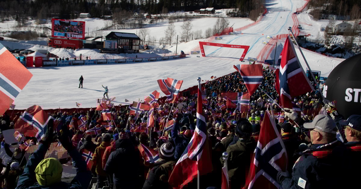 Publikum står tett i en tribune under et skirenn og vifter med norske flagg, med målområdet og løypa synlig i bakgrunnen.
